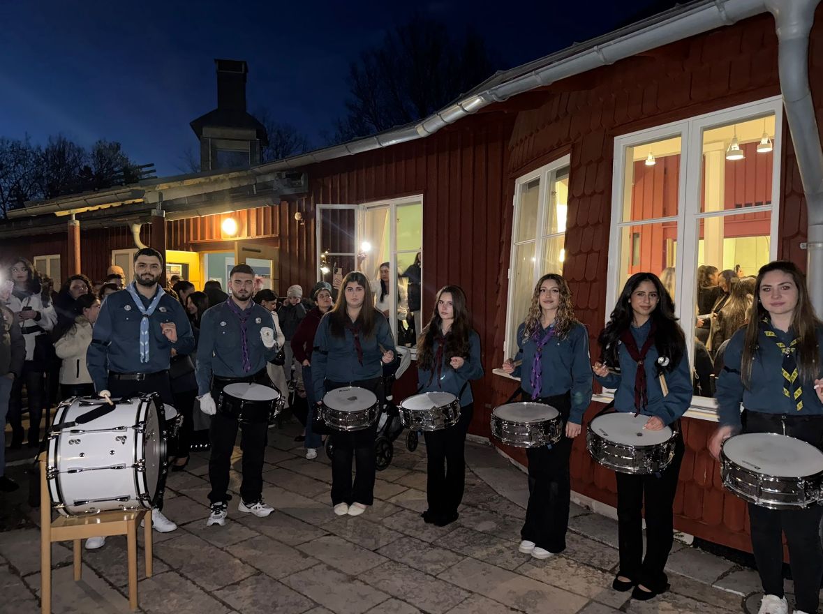 Scouter i uniform spelar trummor utanför ett rött trähus i kvällsmörkret